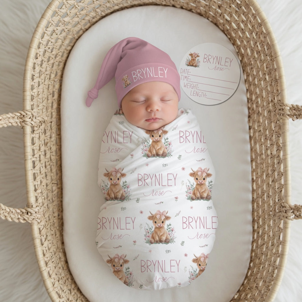 Newborn baby girl swaddled in personalized Highland Cow pink swaddle with matching knotted hat and birth stat sign